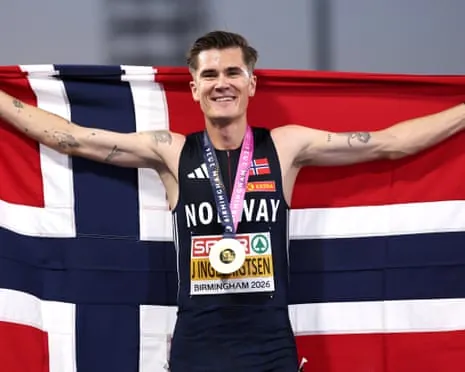 Jakob Ingebrigtsen smiles with a gold medal, holding up the Norwegian flag.